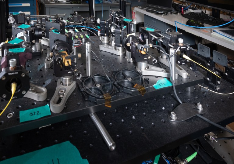 A complex optical experimental setup on a lab table. Various lenses, mirrors, and equipment are mounted on a perforated metal board, with cables and wires connecting components. Small labels with numbers and letters are visible on the apparatus.