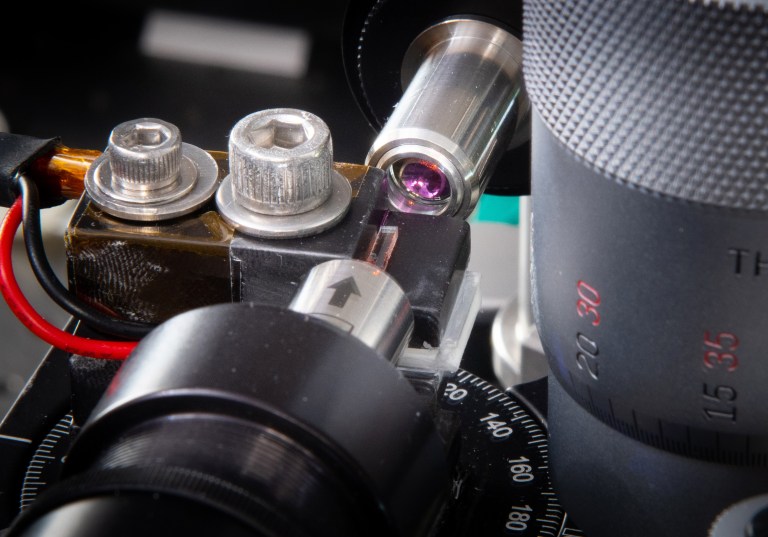 Close-up of a microscope setup, featuring a stage with adjustable knobs and a lens focusing on a specimen. The image shows intricate parts including screws, wires, and a protractor-like dial, indicating precise scientific equipment.