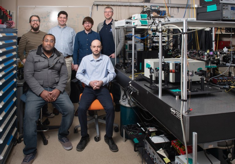 Six men in a laboratory setting, five standing and one seated on an orange chair. They are surrounded by electronic equipment and tools. A whiteboard with various markings is visible in the background.
