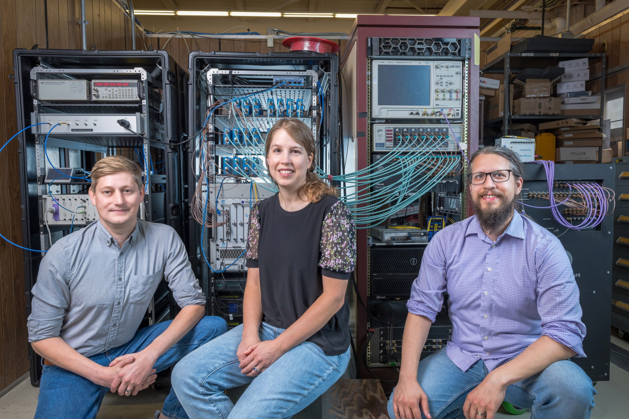 Mike Marsden, Jennifer Downey, and Brian Vyhnalek kneel in front of three electronic racks and one detector unit in a black metal box. In the racks are electronics and optics connected by cables. 