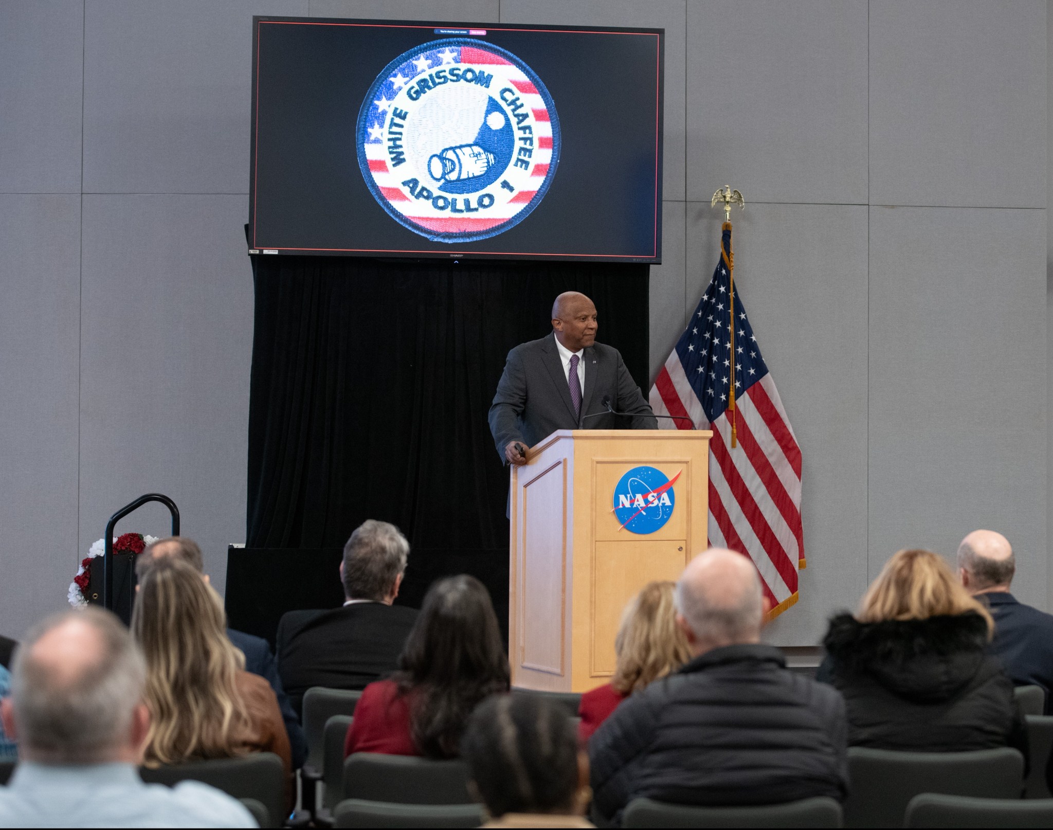 The acting director of NASA’s Kennedy Space Center stands on stage in front of a podium with a NASA emblem and addresses an audience of Glenn employees. The Apollo I mission patch is displayed on a video screen behind him.