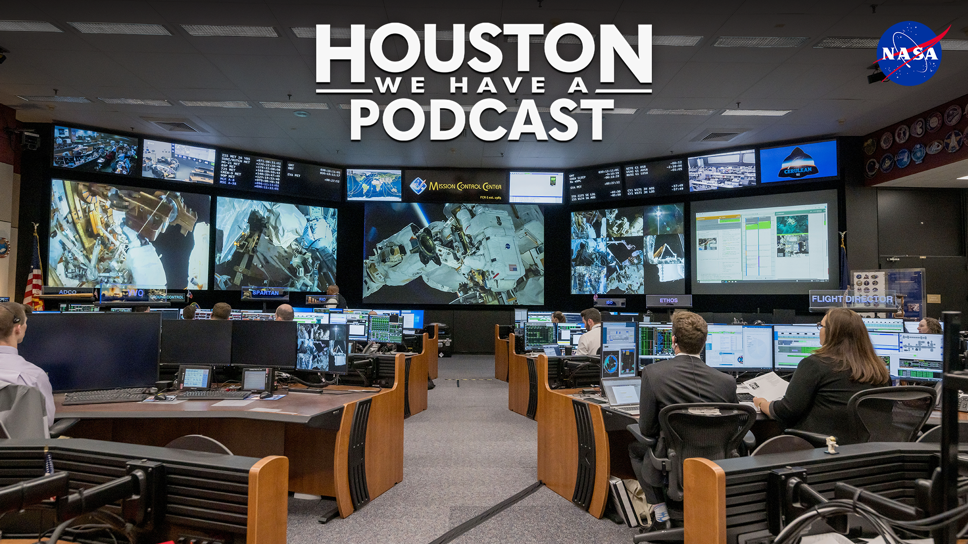 HWHAP Ep. 374. An aisle between desks in ISS Mission Control leads to the front screen showing an image from the Space Station.
