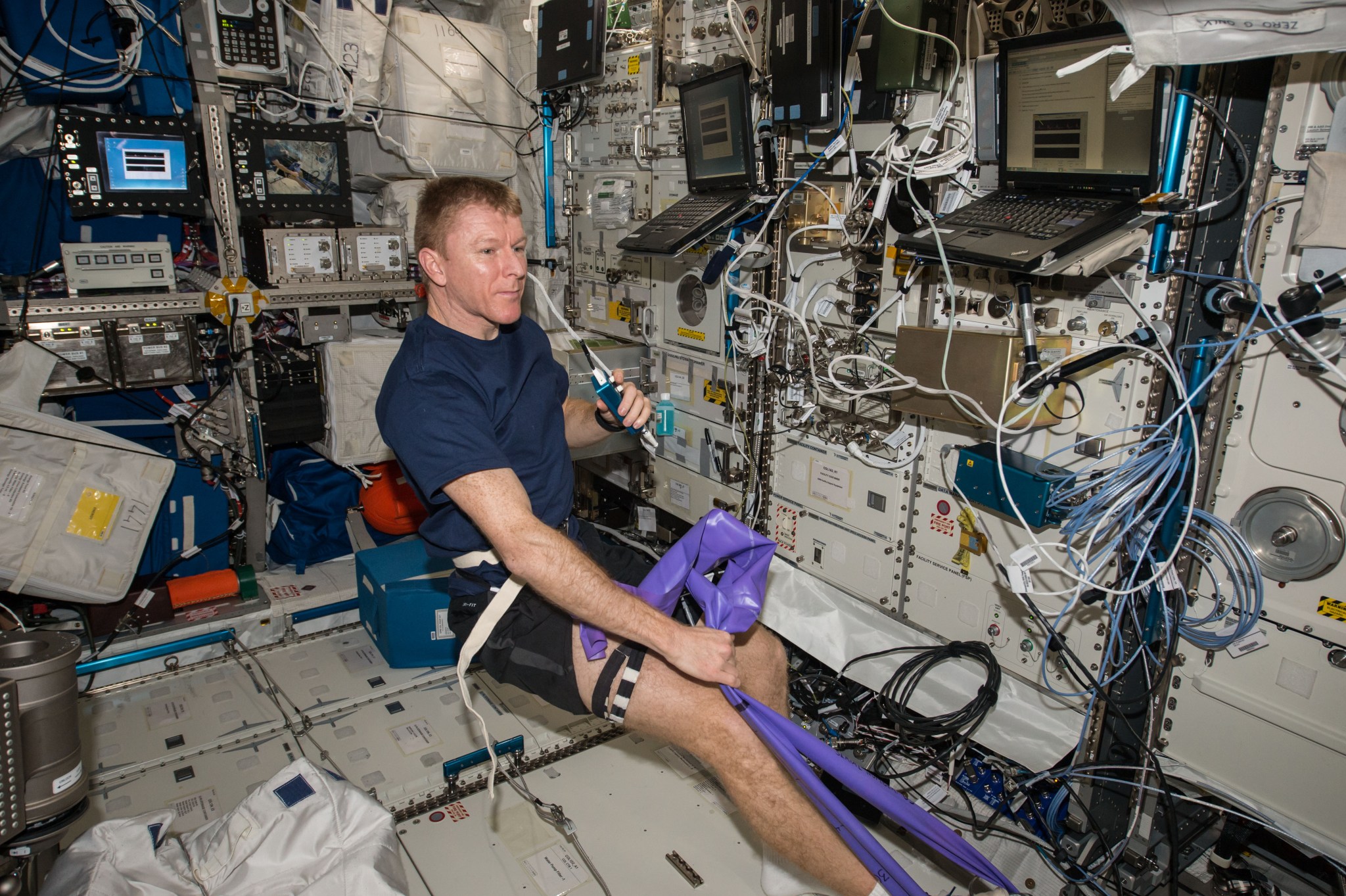 Peake, in a blue t-shirt and black shorts, is using his right hand to pull on a purple resistance band around his right foot. He is holding a small blue microphone in his left hand. Several laptops and multiple cords, wiring, and hardware are visible behind him.