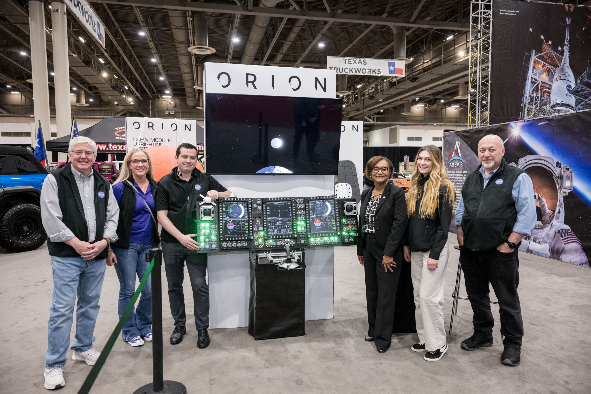 Group photo of six individuals standing in front of an Orion display booth at an exhibit, featuring a digital control panel and a monitor.