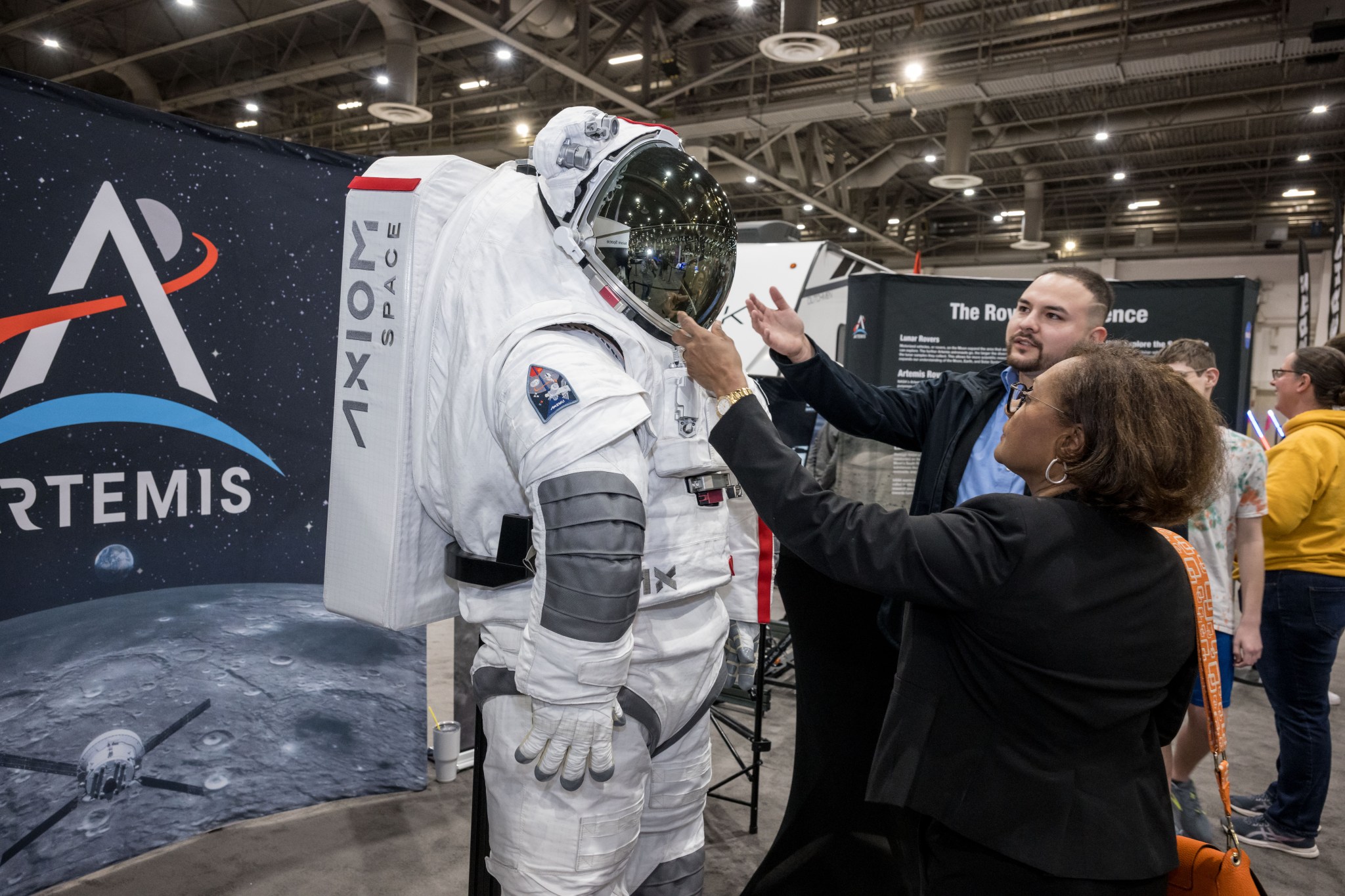 Two individuals examining a detailed Axiom Space spacesuit displayed at an exhibit booth, with an Artemis program banner in the background.