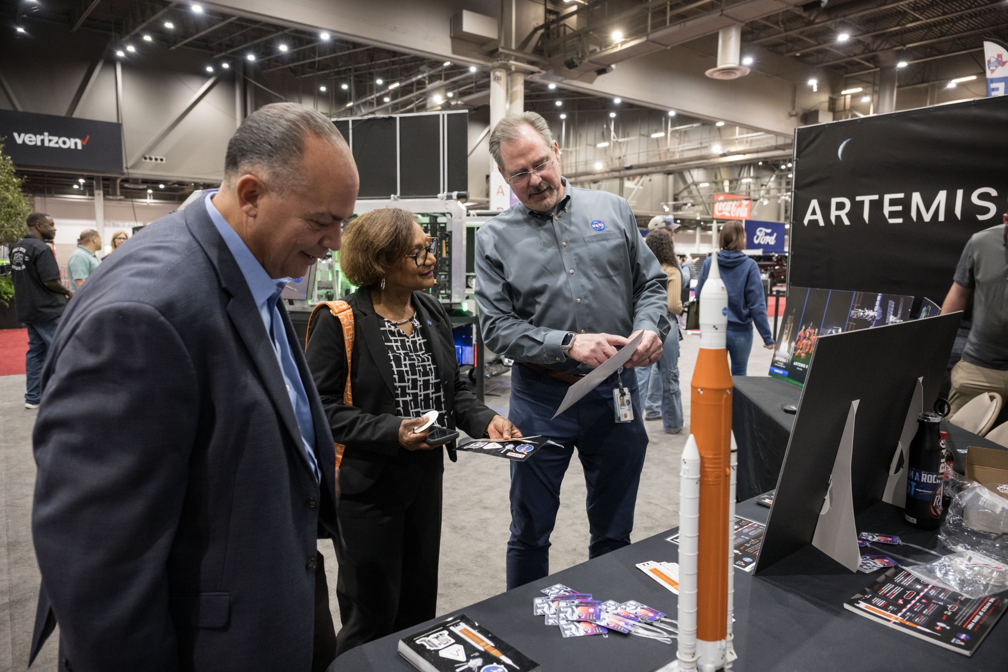 Three individuals engaged in discussion at a display booth featuring NASA’s Artemis campaign.