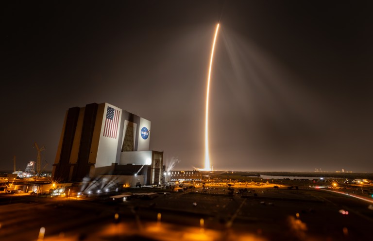 Leaving a golden streak in the sky, a SpaceX Falcon 9 rocket lifts off from NASA's Kennedy Space Center in Florida in the evening. The light from the rocket flames illuminates the misty night sky. NASA Kennedy's Vehicle Assembly Building, a massive rectangular building, is visible at left.
