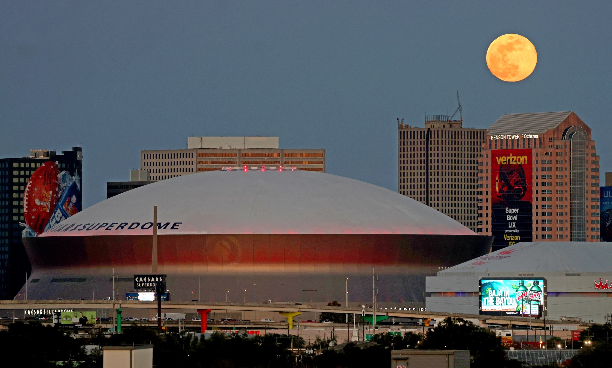 The full moon (top right) rises over buildings in New Orleans, Louisiana, including the Superdome. The Superdome has a white rounded top. Billboards and ads can be seen around and on some of the buildings.