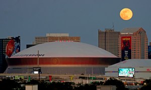 The full moon (top right) rises over buildings in New Orleans, Louisiana, including the Superdome. The Superdome has a white rounded top. Billboards and ads can be seen around and on some of the buildings.