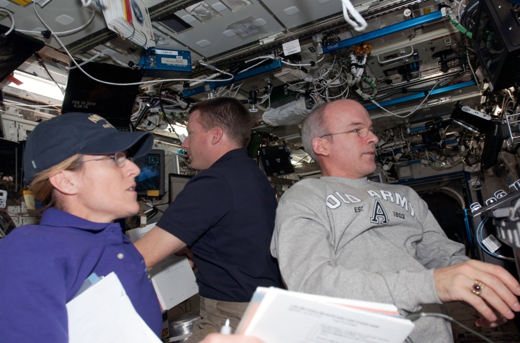 One female and two male astronauts are seen inside the space station operating its robotic arm.