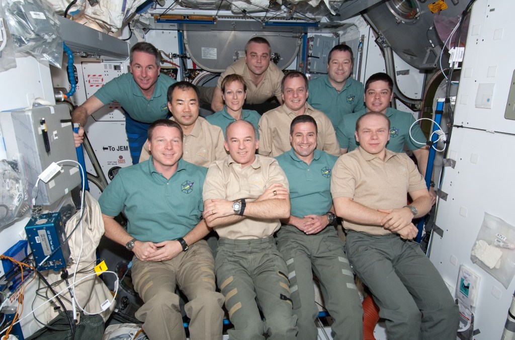 Group photo of 11 astronauts inside the space station, wearing green and gold T-shirts.