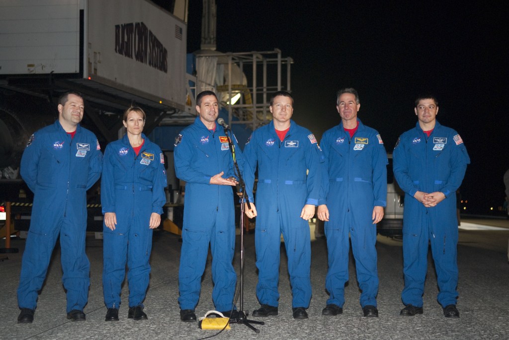 Image of six astronauts wearing blue overalls standing in front of a space shuttle.