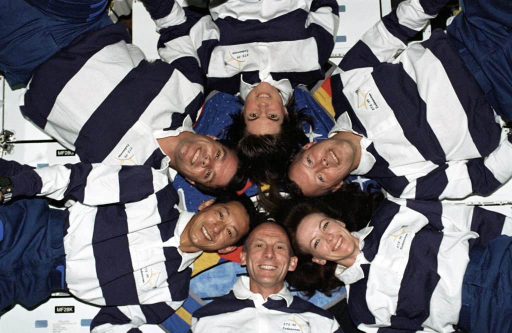 Image of six astronauts wearing blue and white jerseys, floating in a circle with their heads toward the center of the circle.
