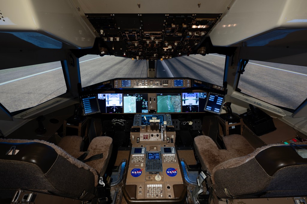 The inside of an airplane cockpit simulator with two seats and a display of various aircraft system information across large screens at the front of the plane with large windscreens and a pilot control panel above.