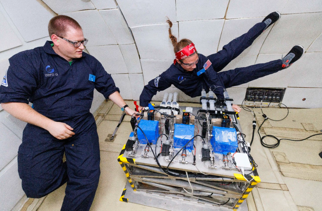 Two researchers wearing blue jumpsuits float in reduced gravity while monitoring their experiment that includes three blue containers.