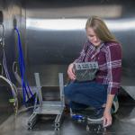 A woman wearing a flannel shirt and jeans kneels on the ground while holding a large metal device. Next to her is a metal apparatus with blue and white cables feeding through holes in the wall.