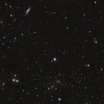 A sea of galaxies of many different shapes and sizes, oriented in all directions and many with spiral arms visible. A large, nearby face-on spiral galaxy draws attention at the bottom center of the image. Two bright stars in the center and to the left are seen with prominent diffraction spikes.