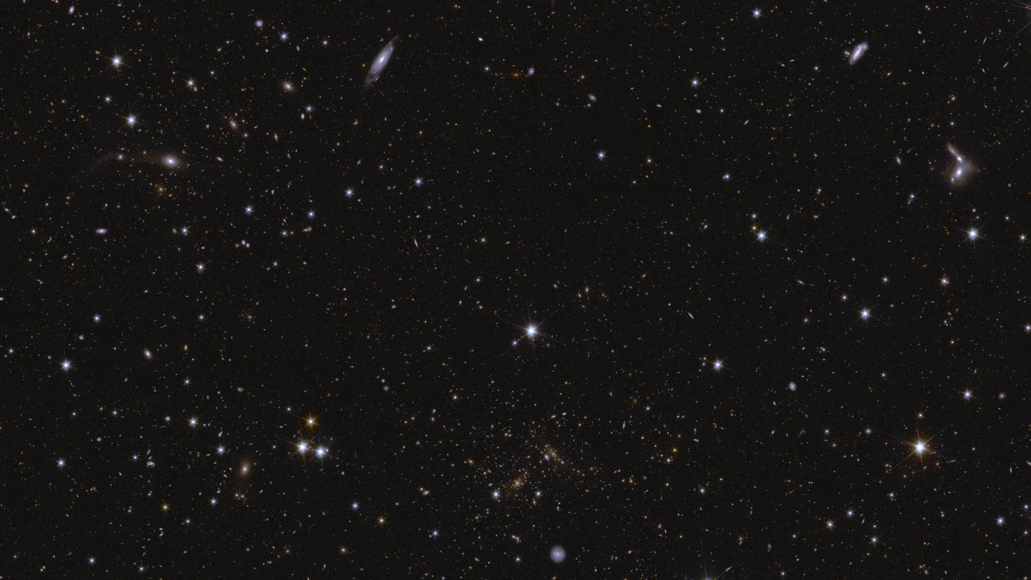 A sea of galaxies of many different shapes and sizes, oriented in all directions and many with spiral arms visible. A large, nearby face-on spiral galaxy draws attention at the bottom center of the image. Two bright stars in the center and to the left are seen with prominent diffraction spikes.