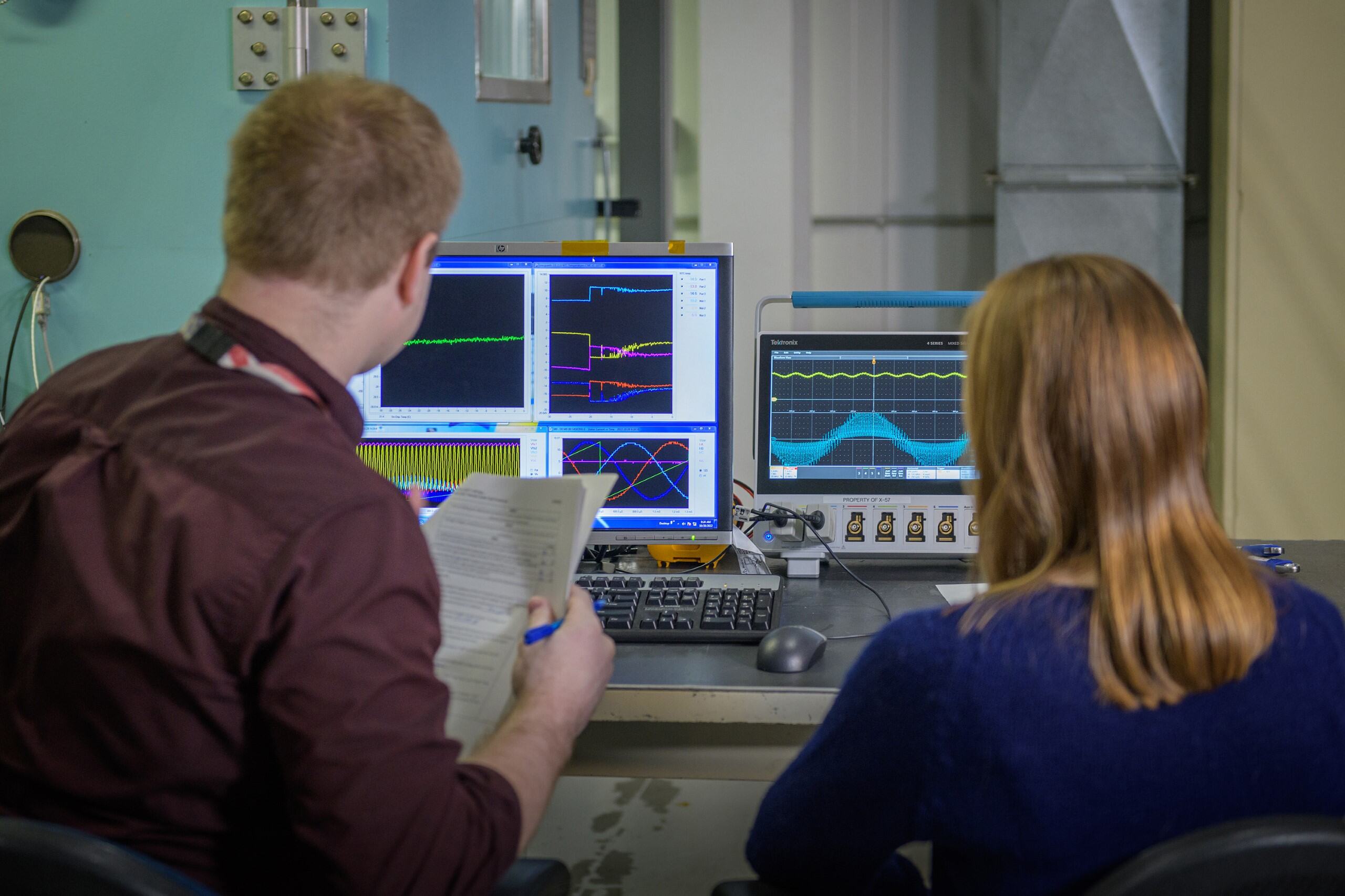 A man holding a packet of paper and a woman face away from the camera looking at various testing data displayed on computer screens.