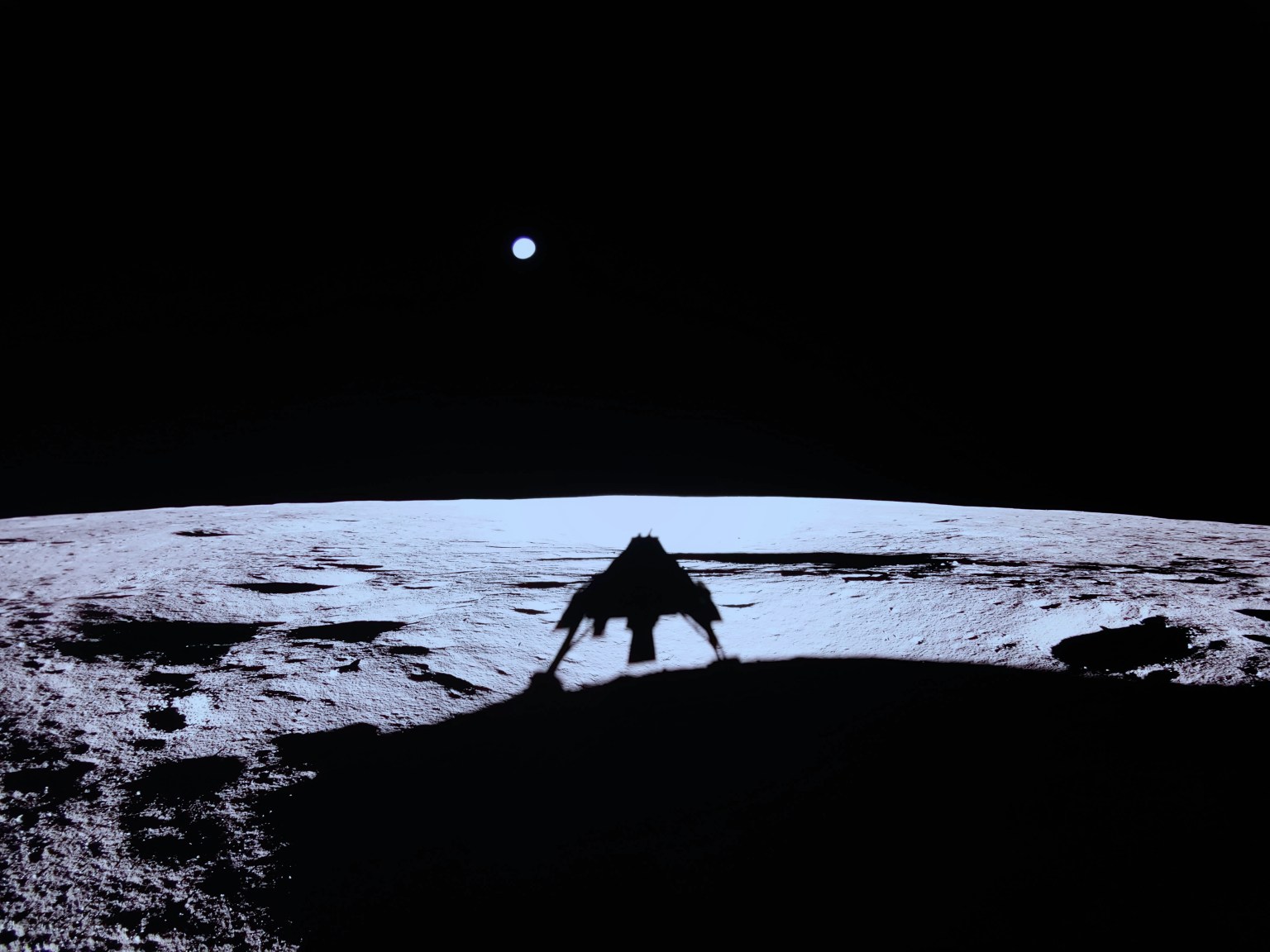 Earth is a small pale circle in the sky in this image taken on the Moon by a camera on @Firefly’s Blue Ghost lunar lander. The Moon’s gray surface sharply contrasts with the blackness of space. The lander’s shadow can be seen on the ground.
