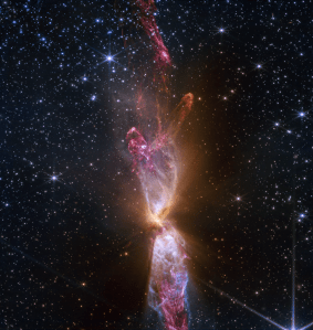At the center is a thin vertical cloud known as Lynds 483 that is shaped like an hourglass with irregular edges. At lower center are two discrete bright white, tiny blobs of light that have raced away from the hidden central stars. The top lobe shows a more prominent orange U-shape. Orange bleeds into light purple, and brighter pink at its edges. Some background stars are visible through sections of this lobe. Higher up, there is an orange arc. Some brighter pink material extends to the top edges near the center. In the lower lobe, less orange is visible. More opaque light purple is in its top third, rippling out into semi-transparent blues and pinks. The lower lobe has more texture. V-shapes left and right of the lobes are darkest, and the background stars in these areas appear orange. Elsewhere, the black background of space is clearer, speckled with tiny white stars and faint orange galaxies.