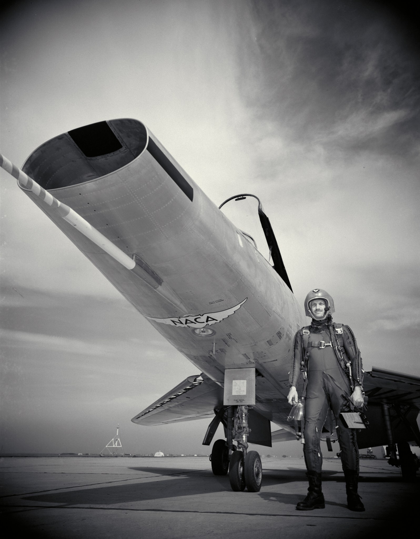 George Cooper, a test pilot for the National Advisory Committee for Aeronautics stands next to a North American F-100 airplane at what is now NASA’s Ames Research Center. Cooper wears a helmet and a tight jumpsuit with a harness. "NACA" is visible on the underside of the plane.