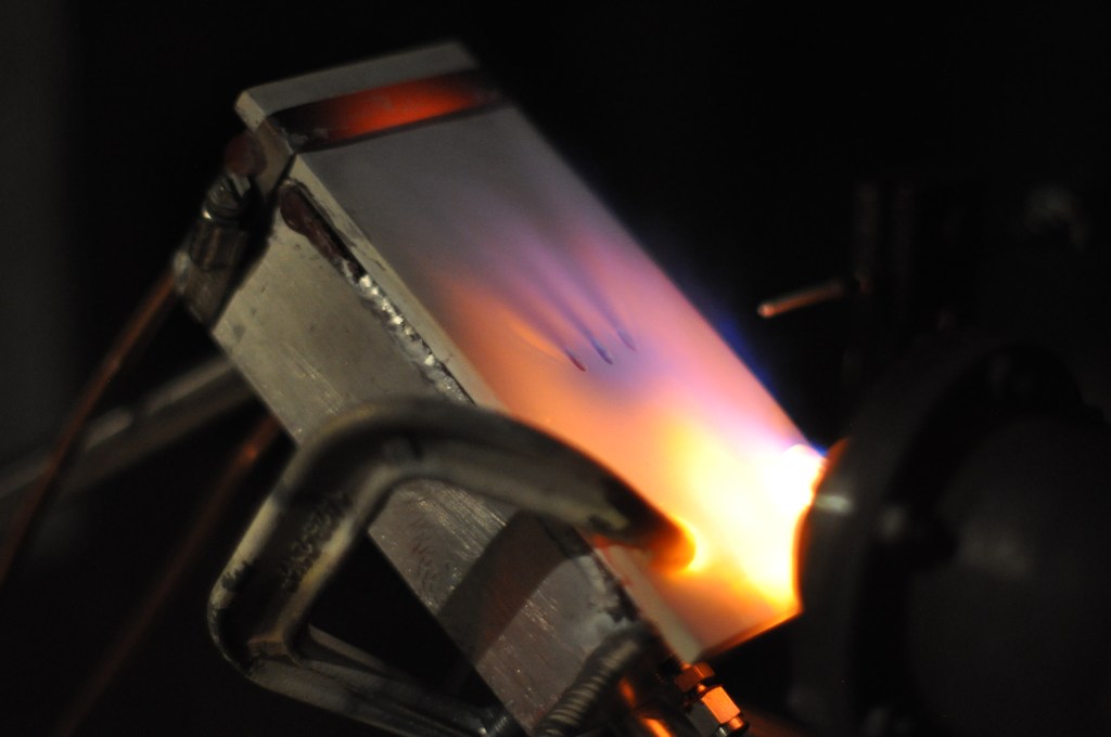 A metal plate is being heated with an orange flame in a workshop setting. The metal plate is held in place by clamps.
