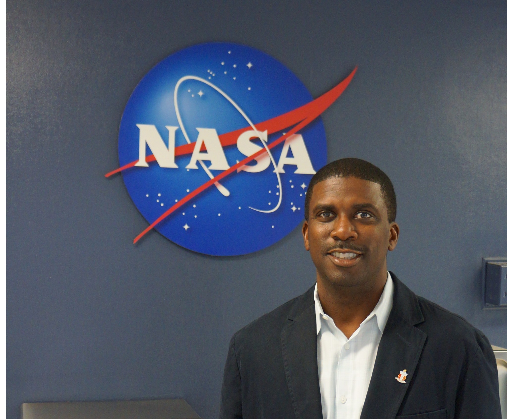 A man stands and smiles in front of a NASA meatball. He is wearing a white button down and black blazer.