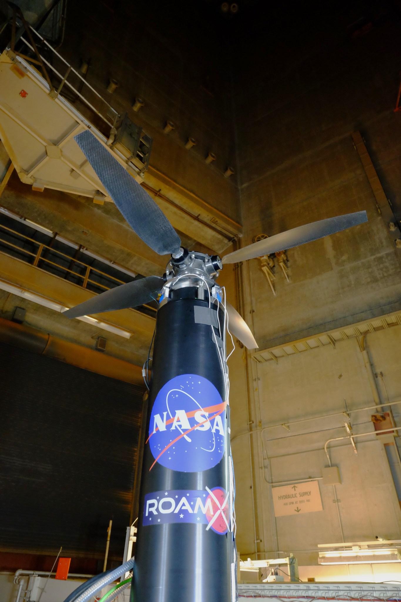 A vertical rotorcraft with four large carbon-fiber rotor blades, featuring NASA and ROAMX logos, inside the Planetary Aeolian Laboratory (PAL) at NASA Ames Research Center. The rotorcraft, part of the ROAMX project, was tested under simulated Martian atmospheric conditions to optimize helicopter performance for future Mars missions.
