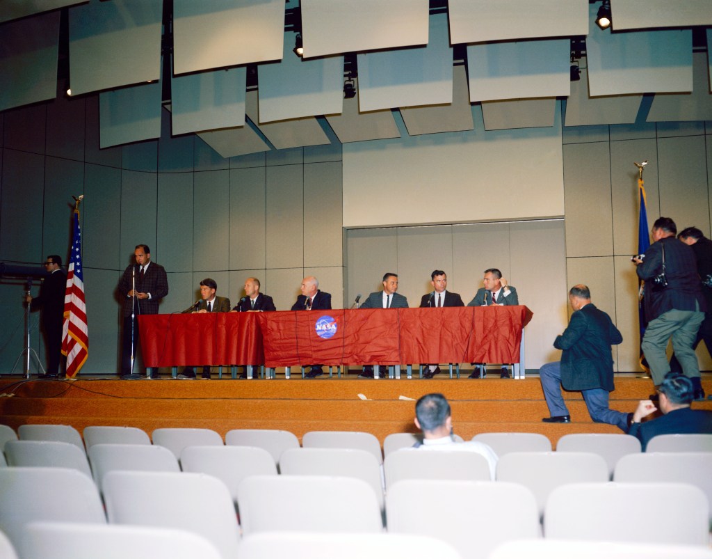Image of six men sitting behind a long dais on the stage of an auditorium.