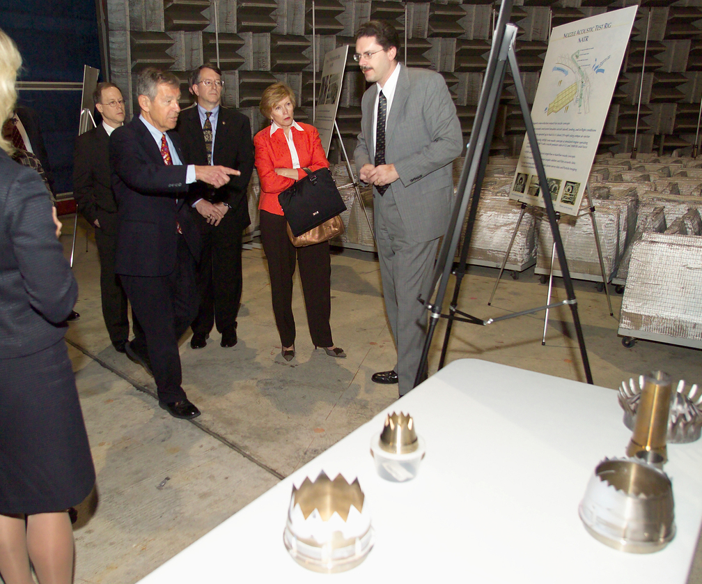 A NASA researcher presents information on a poster to five people inside a NASA acoustic facility. They wear business professional clothing. A man in the front of the group points to the poster. On a white table in the foreground are small, circular, silver-colored chevron nozzles with jagged edges.