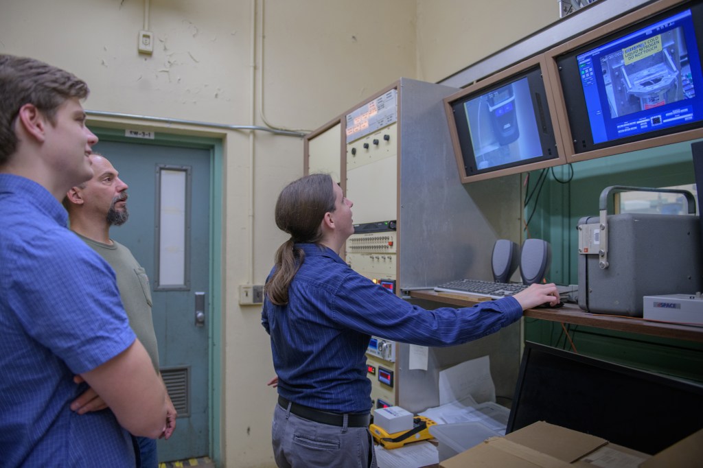 Three men stand inside a test facility looking upwards at large monitors on the right side of the wall.