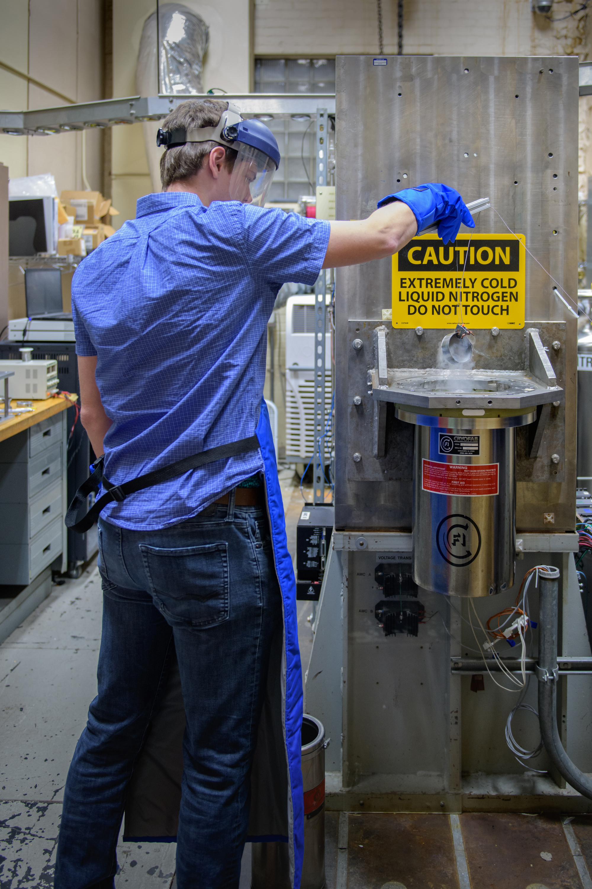 A man wearing a blue shirt, jeans, and an apron dips a test article into a metal cylinder. The sign above the rig includes the words “Caution. Extremely cold liquid nitrogen. Do not touch.”