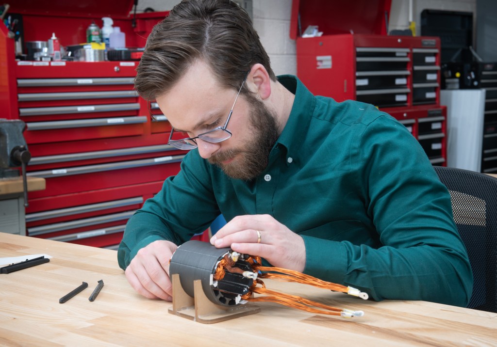 A man wearing a dark green button-down shirt sits at a table and feeds copper-colored wires into a cylindrical metal object. Behind him are red tool storage containers.