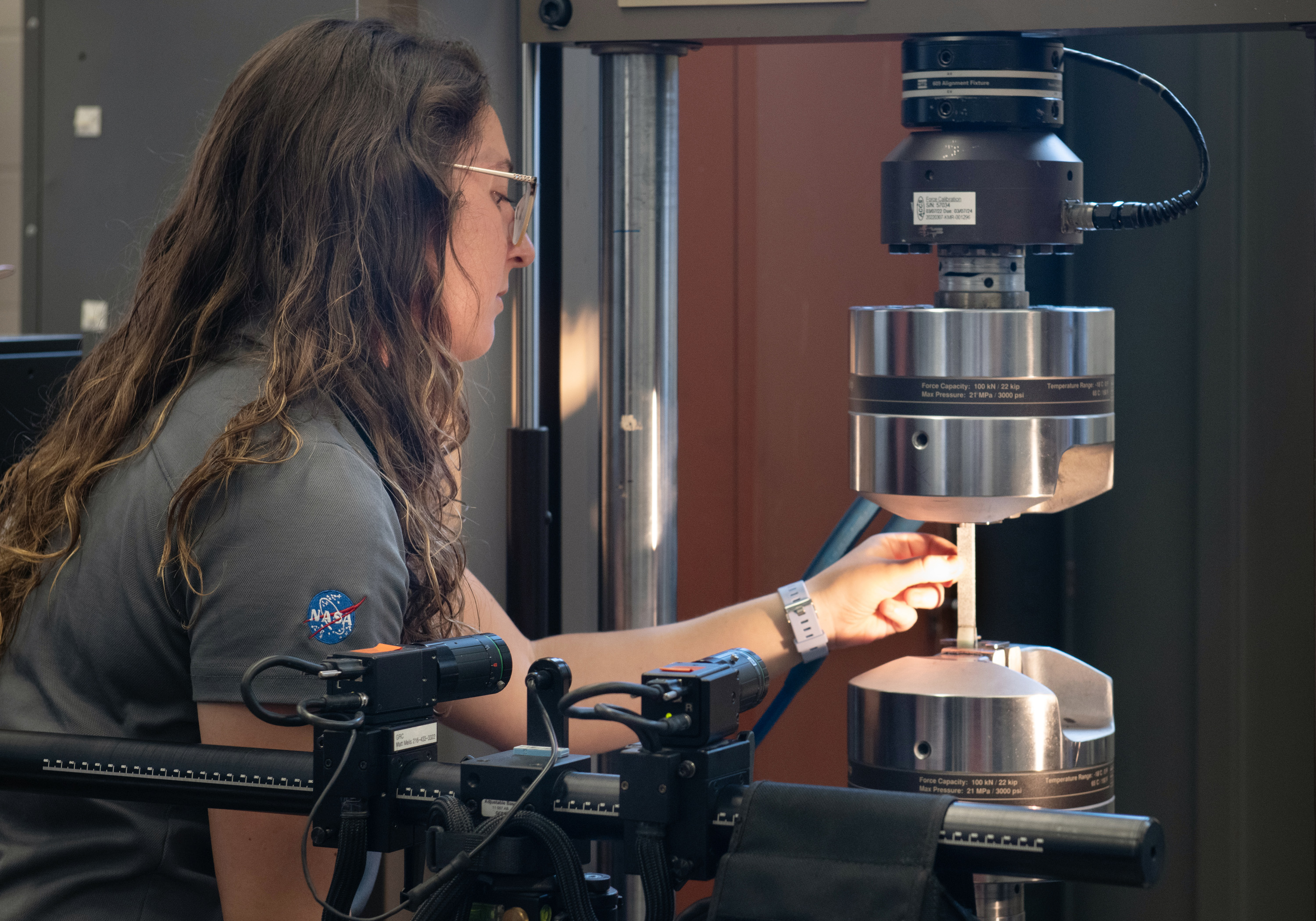 A woman with long brown hair examines a thin piece of material wedged in the middle of two metal testing devices.