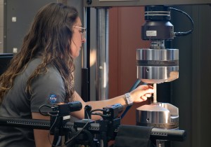 A woman with long brown hair examines a thin piece of material wedged in the middle of two metal testing devices.
