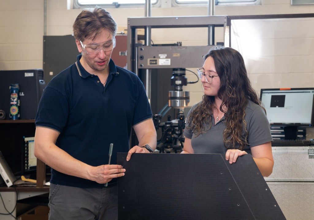 A man and a woman stand next to each other as they examine a large piece of composite material, with various hardware and testing machinery in the background.