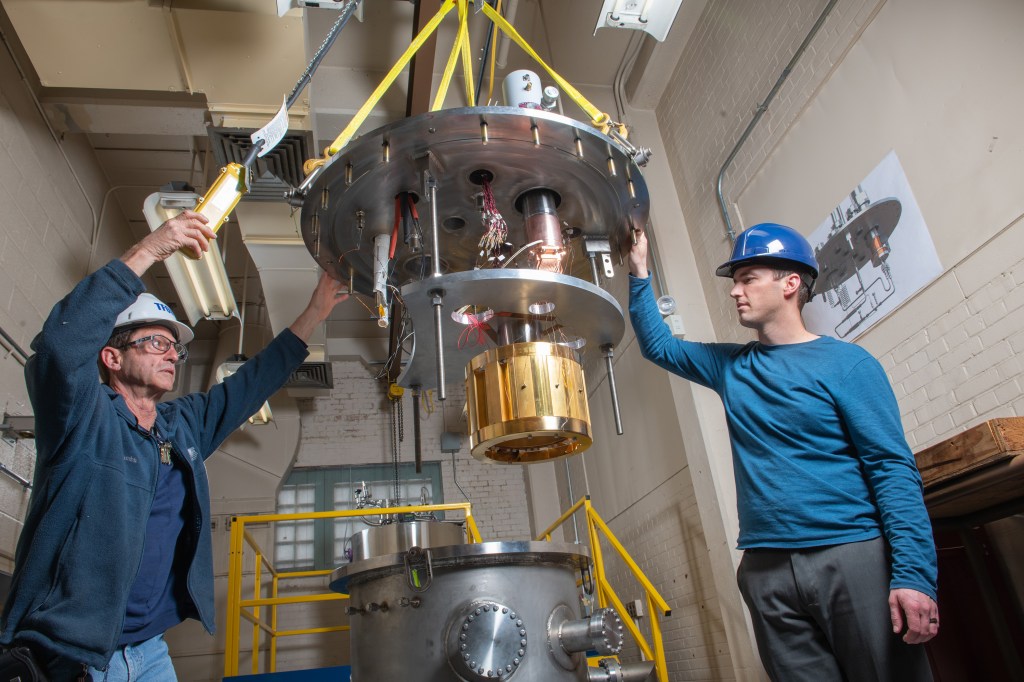 Two men wearing hard hats help hoist a piece of hardware above a circular cryogenic testing rig.