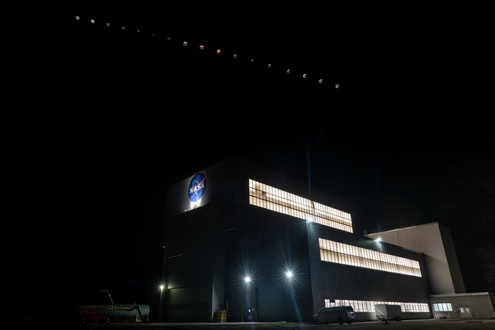 A time-lapse image shows the phases of the lunar eclipse, including the blood moon, on March 14, 2025. The Moon travels through the sky above a large building with the NASA meatball logo on it. There are a few spotlights on the building, and lights illuminate the large rectangular windows along the length of the building on several floors. The image is otherwise dark.