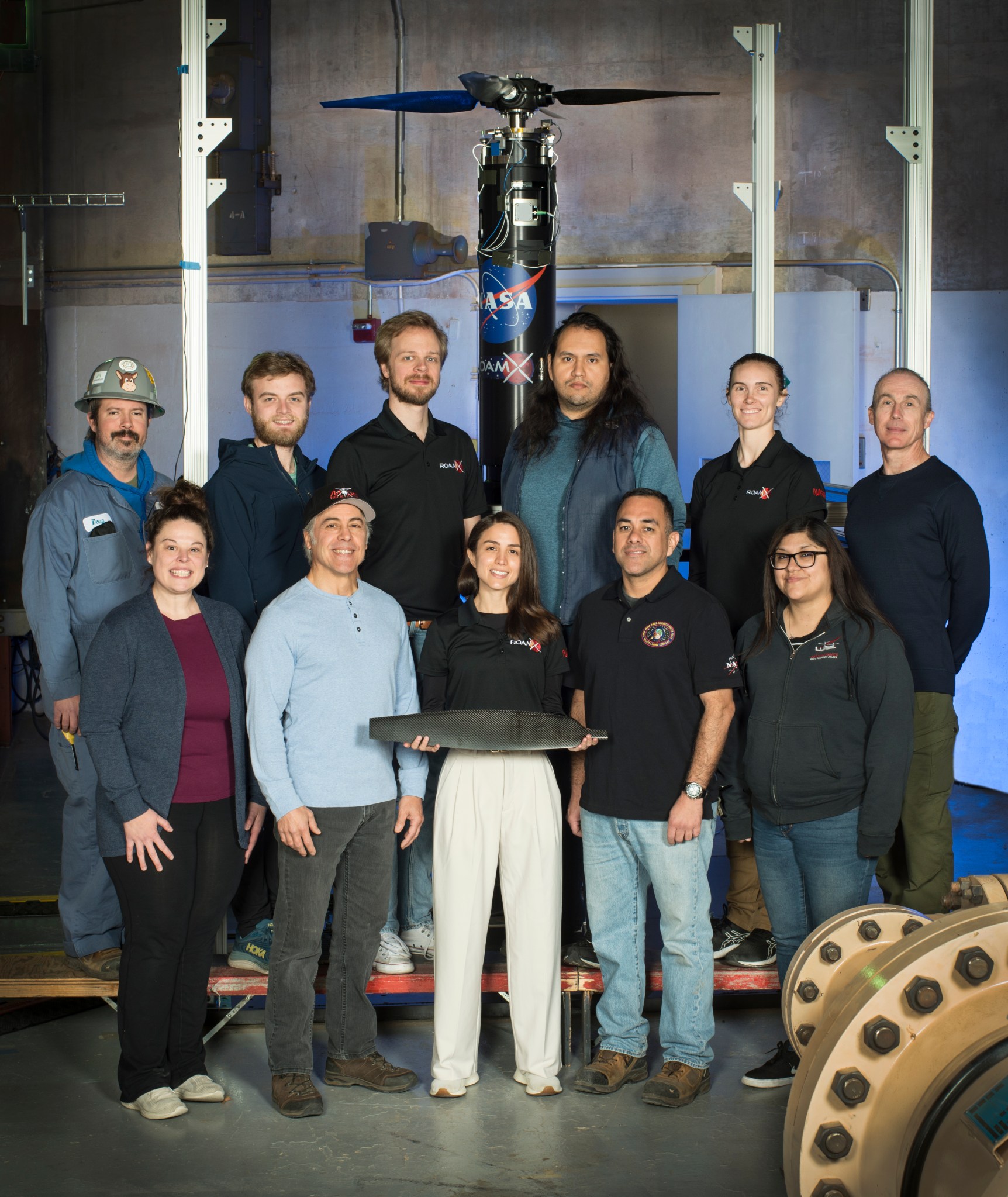A group of NASA researchers and engineers pose in front of the ROAMX rotorcraft inside the Planetary Aeolian Laboratory (PAL) at NASA Ames Research Center. The team, consisting of Douglas Gray, Stephen Wright, Witold Koning, Antonio Cervantes, Haley Cummings, Alex Sheikman, Natasha Schatzman, Charles Cornelison, Natalia Perez, Alfredo Perez, and Michelle Dominguez. They tested helicopter blades optimized for Mars, with a researcher (Natalia Perez) in the center holding a rotor blade