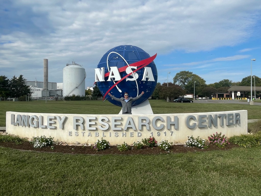 An aerospace engineer raises his arms joyfully while standing in front of the NASA meatball and sign that reads NASA Langley Research Center.