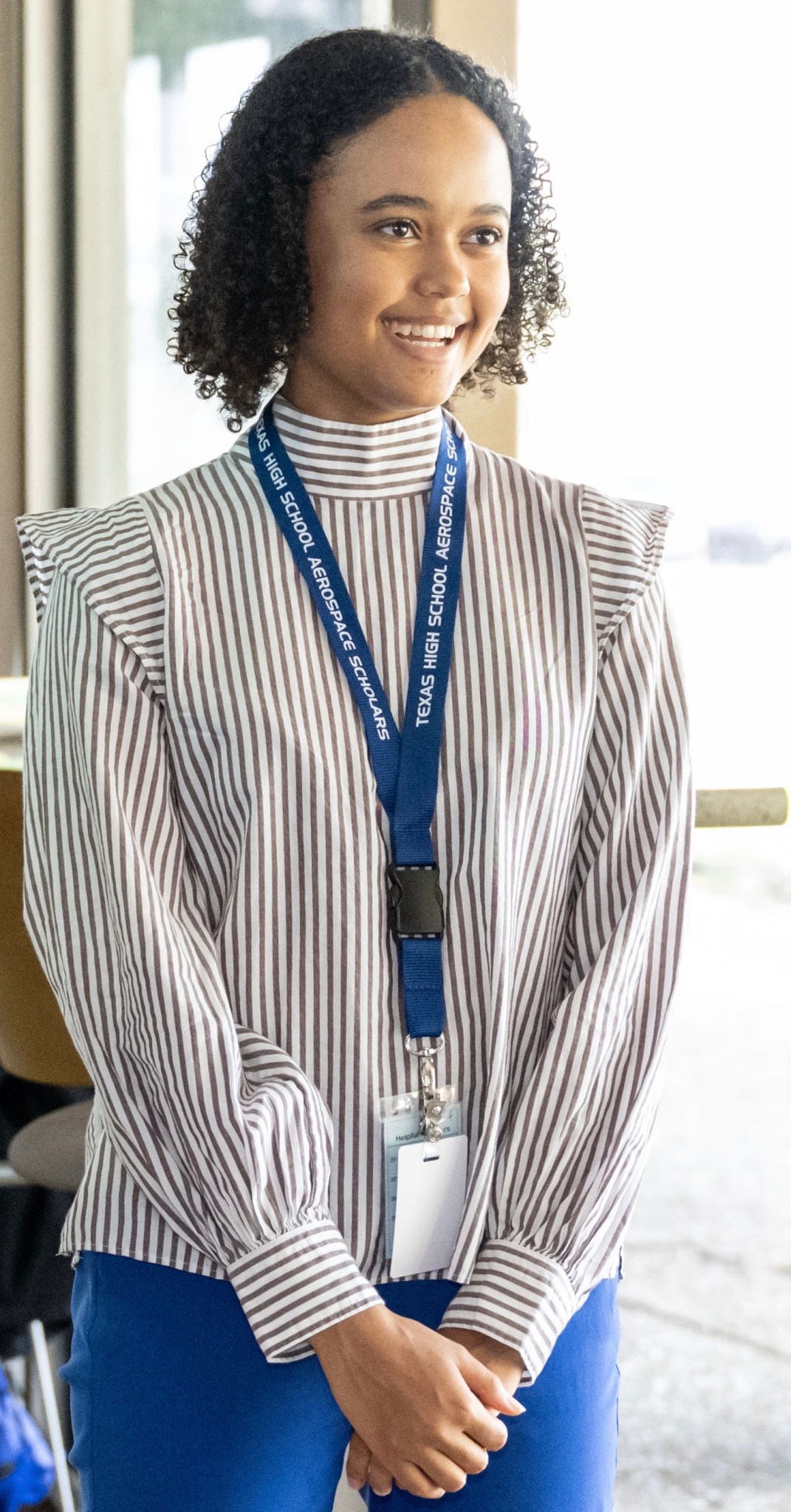 A girl smiles in a striped blouse and blue pants. She is wearing a blue lanyard. A window is behind her and it is a bright sunny day.
