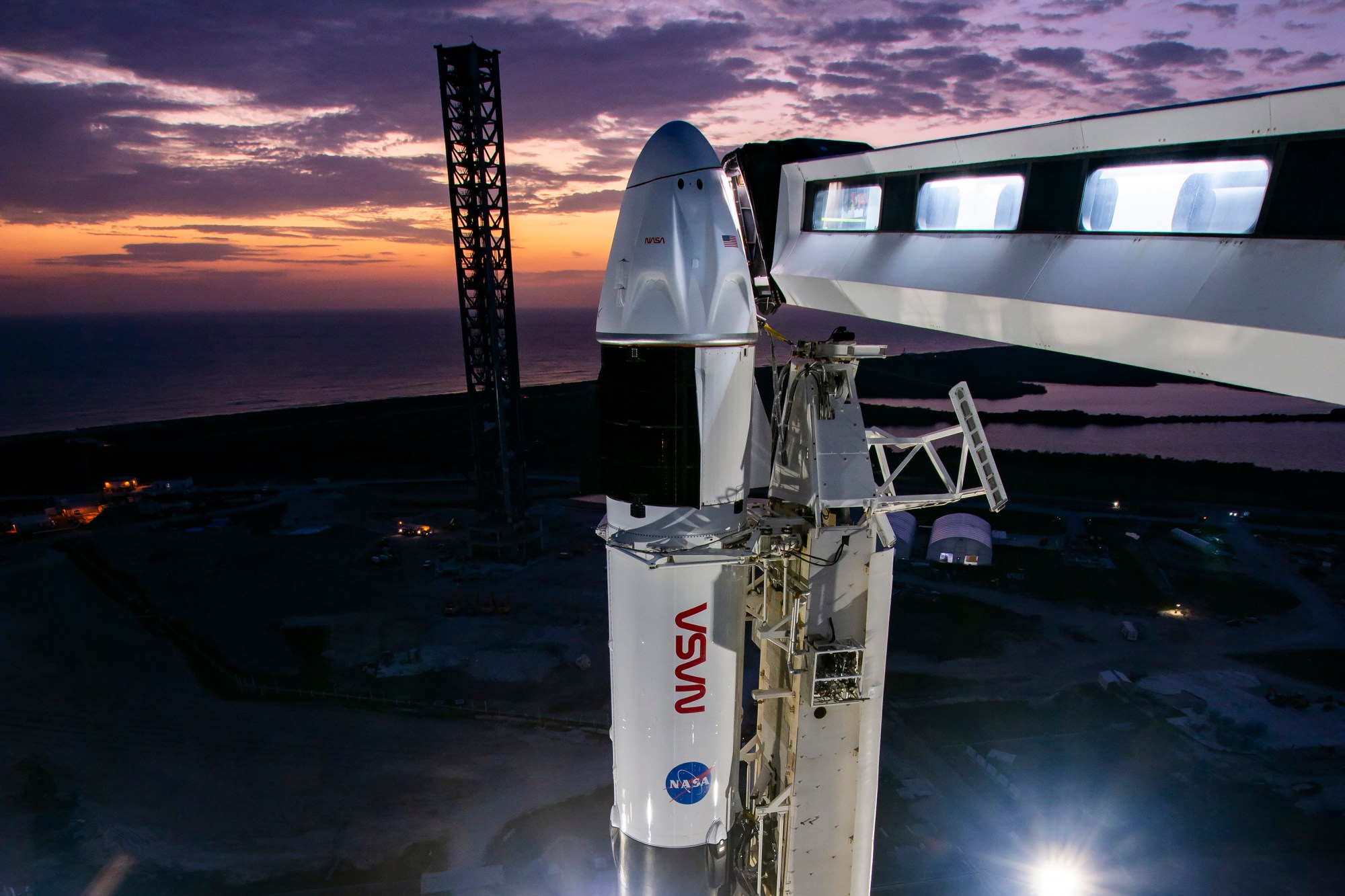 The upper portion of a rocket with a spacecraft on top are visible in this picture. Both NASA logos are visible - the "worm" and the "meatball." A white bridge with several rectangular windows connects to the Dragon spacecraft. The Sun is rising in the background, brightening the sky slightly with an orange glow. The ground below is dark.