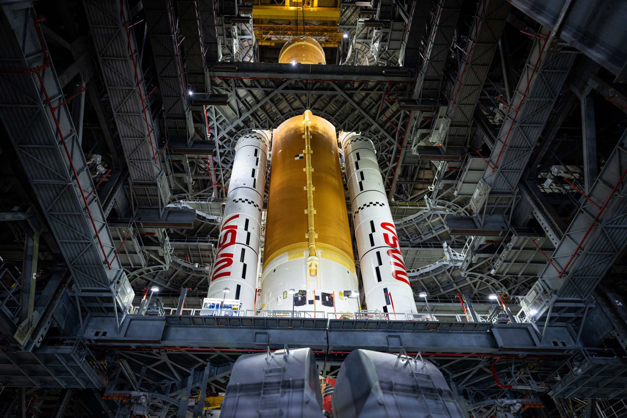 The Artemis II core stage (a butterscotch orange cylinder in the middle) and solid rocket boosters (the white cylinders, branded with the NASA "worm" logo on each side of the core stage) are joined together by teams inside the Vehicle Assembly Building (VAB). The view is from the ground, looking far upward, to show the full height of the core stage - 212 feet. The inside of the VAB has many platforms and catwalks around the center of the building, which is open.