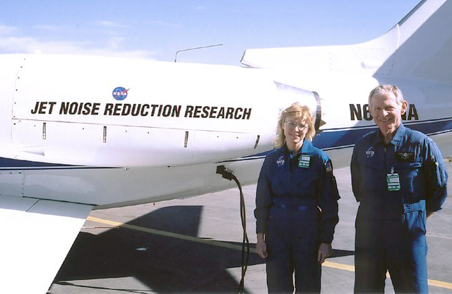 A man and a woman wearing blue NASA flight suits stand in front of the tail of a white NASA airplane with a blue stripe along its side, a small NASA logo, and text that says, “JET NOISE REDUCTION RESEARCH.”