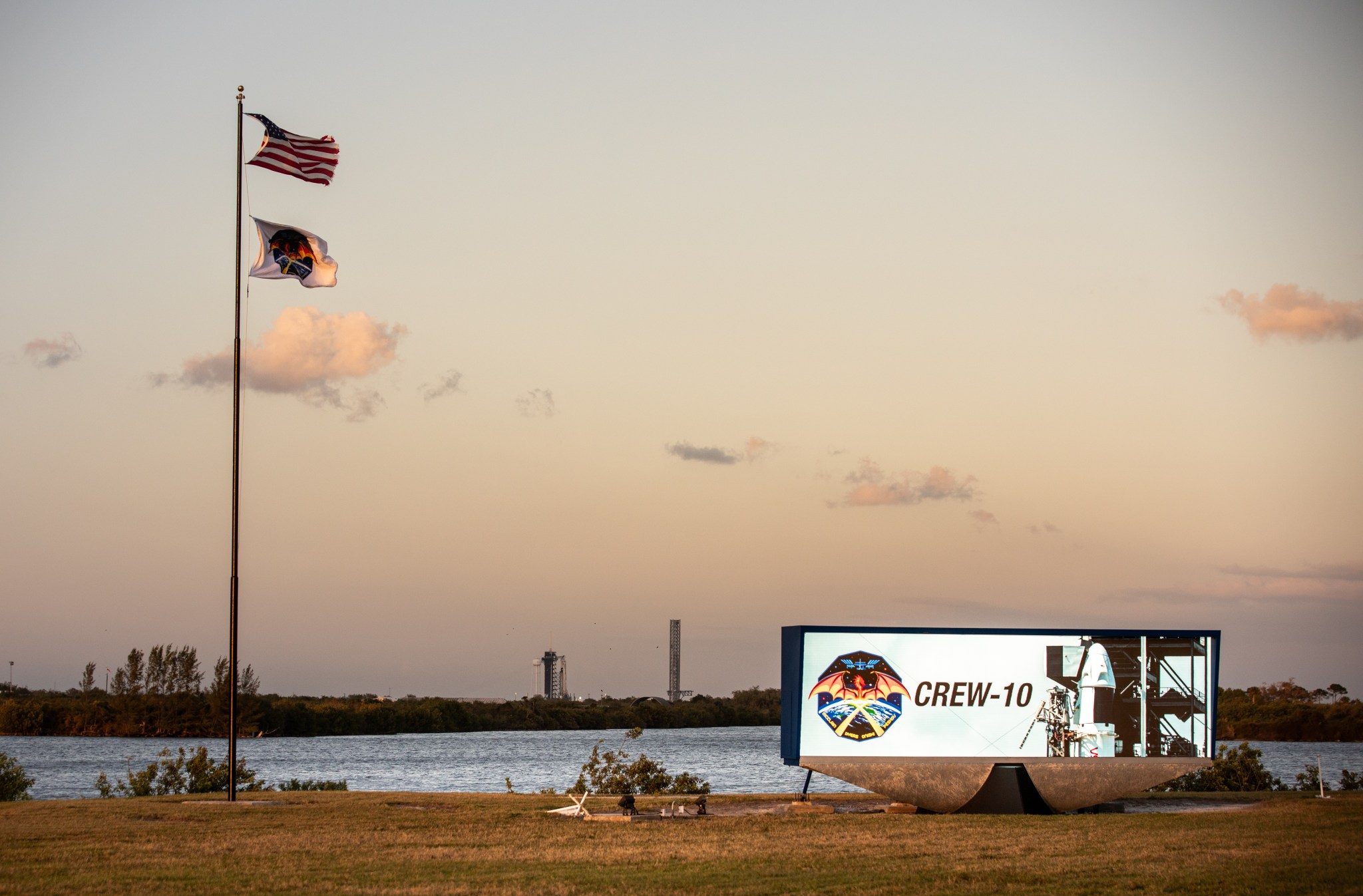 Image shows Crew-10 in monitor, along with two flags, overlooking Launch Complex 39A