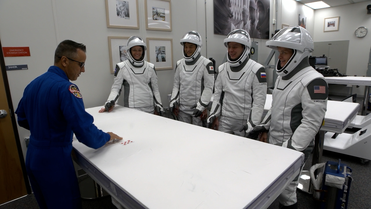 Image shows three astronauts and a cosmosnaut standing at table with cards.