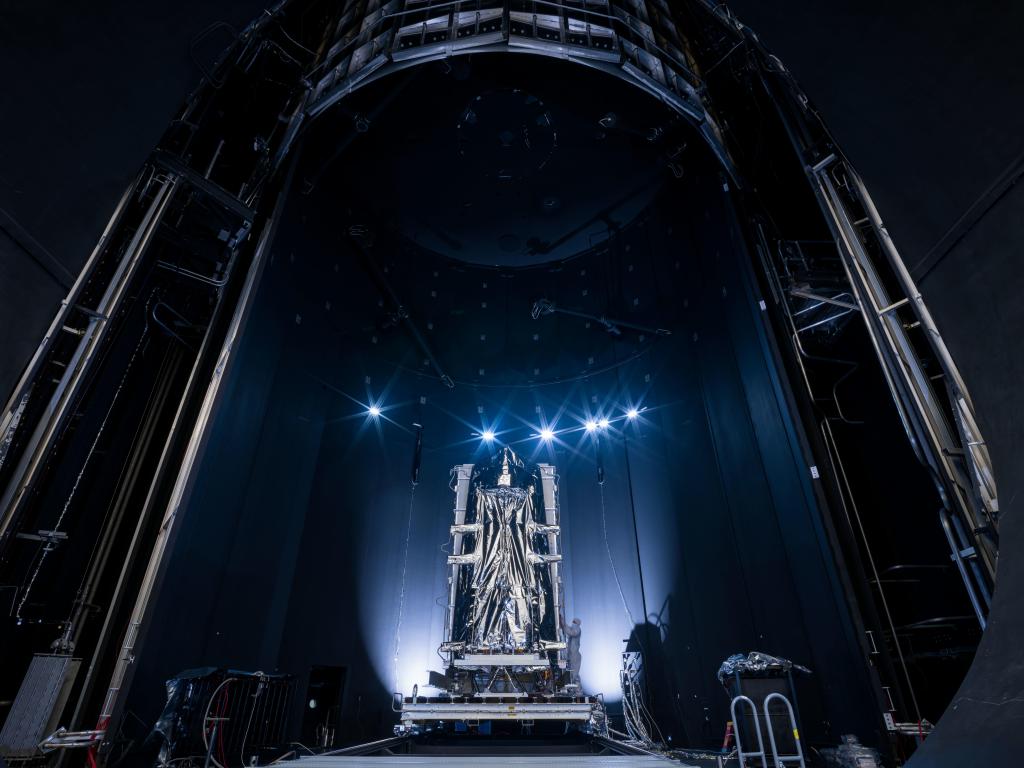 A picture of a massive, silver, statuesque piece of hardware inside a cavernous testing chamber. The image is mostly dark, with an illuminated section in the lower center half of the picture. The hardware has two large, vertical silver metal posts on either side of it, and two silver metal posts that cross horizontally between them. In the center of those posts is a large portion of silver thermal blanketing that is gathered toward the middle. White lights shine upward from the base of either side of the hardware. A line of six, small, white lights with a blueish starburst effect crown the hardware.