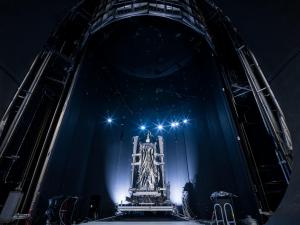 A picture of a massive, silver, statuesque piece of hardware inside a cavernous testing chamber. The image is mostly dark, with an illuminated section in the lower center half of the picture. The hardware has two large, vertical silver metal posts on either side of it, and two silver metal posts that cross horizontally between them. In the center of those posts is a large portion of silver thermal blanketing that is gathered toward the middle. White lights shine upward from the base of either side of the hardware. A line of six, small, white lights with a blueish starburst effect crown the hardware.