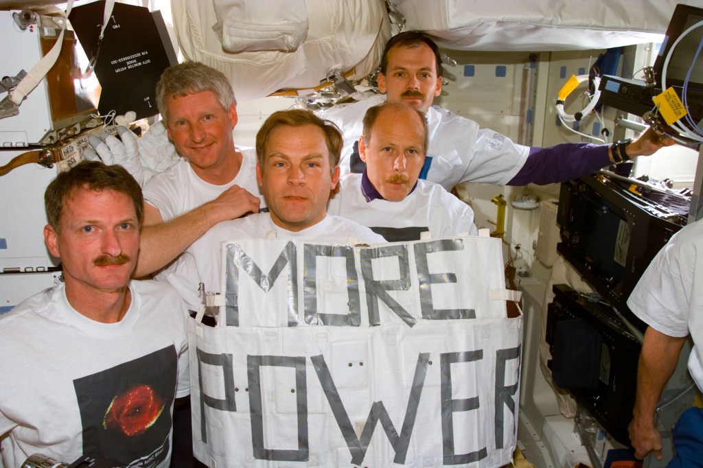 STS-82 crew members prepare for portrait in the middeck with "More Power" banner and Hubble shirts